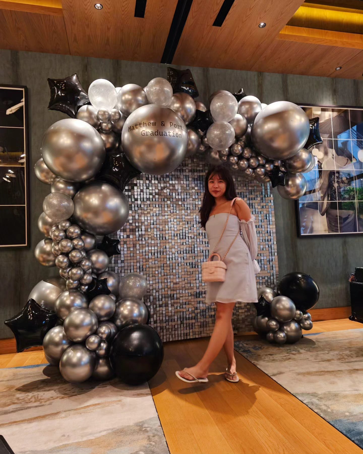 A balloon backdrop featuring a mix of silver and black balloons with a shimmering, reflective surface in a room, with a woman standing in front of the backdrop.